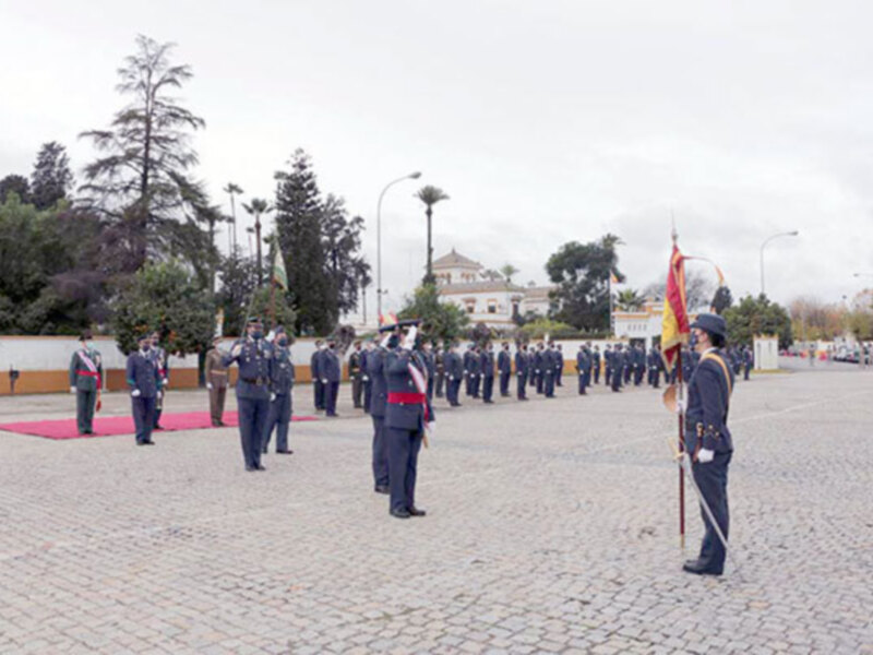 Desfile militar en una plaza pavimentada con uniformes y banderas, con edificios y árboles al fondo.