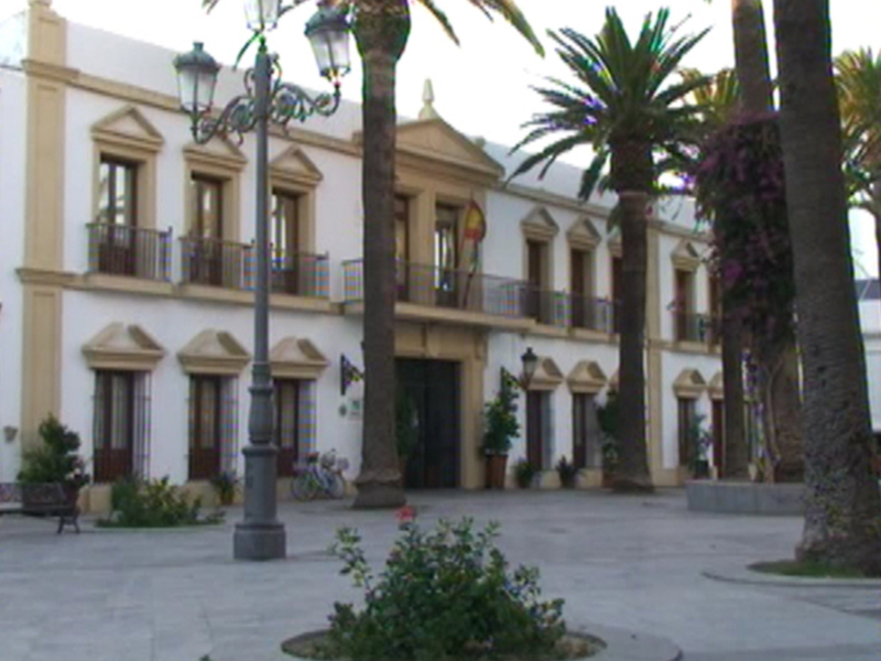 Clásico edificio blanco con balcones y bandera española, rodeado de palmeras y jardines en un entorno urbano.
