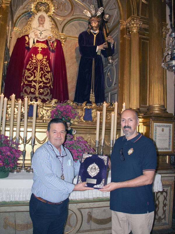 Dos hombres posan frente a una imagen religiosa, sosteniendo un trofeo en un entorno eclesiástico.