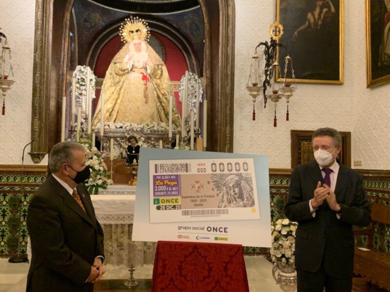 En una estancia decorada con mosaicos y un altar central, dos hombres en trajes de negocios se encuentran frente a una gran tarjeta publicitaria. La imagen muestra un evento de negocios o presentación, posiblemente relacionado con el grupo social ONCE.