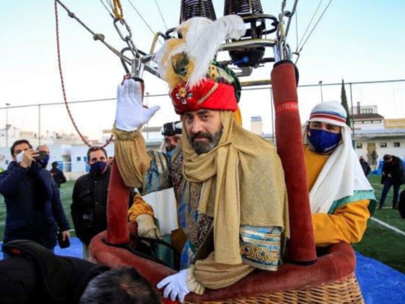 Personaje vestido como rey mago en globo aerostático, saludando durante un evento festivo.