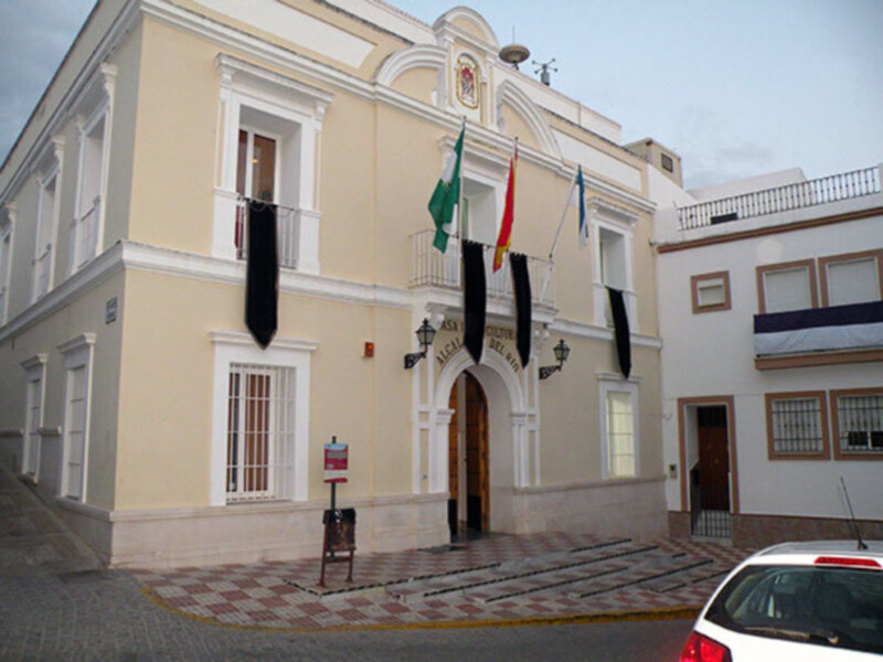 Edificio municipal con banderas y entrada principal en una plaza adoquinada.