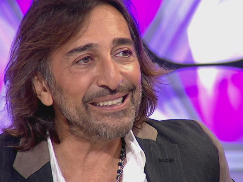 Un hombre con cabello largo y barba sonriendo en un fondo de color púrpura y blanco.