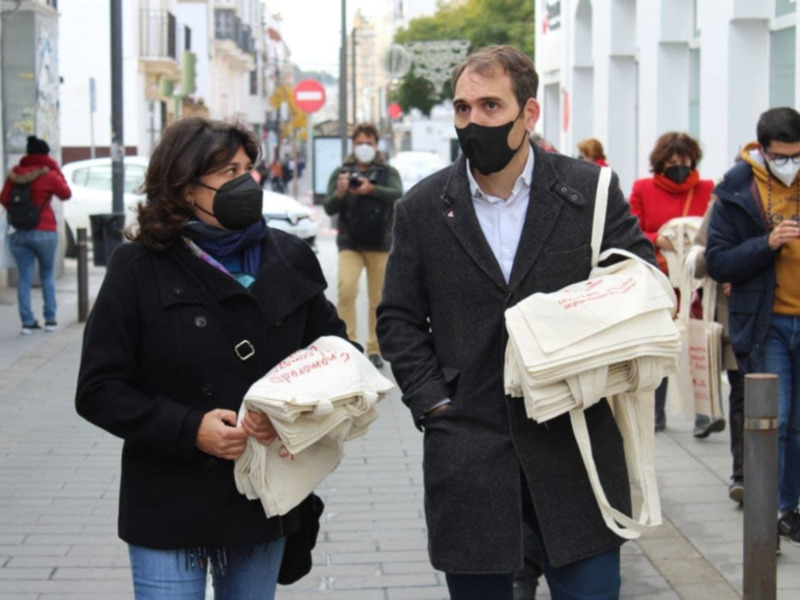 Dos personas caminan por una calle urbana, ambas con mascarillas y bolsas de tela. El ambiente sugiere un día de compras o actividades cotidianas.
