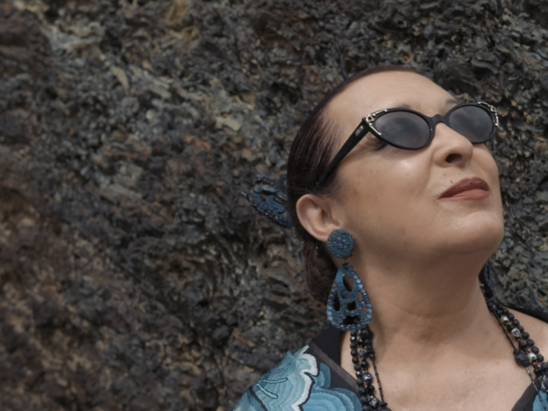 Mujer con gafas de sol y pendientes grandes, mirando hacia arriba con una expresión serena contra un fondo de roca volcánica.