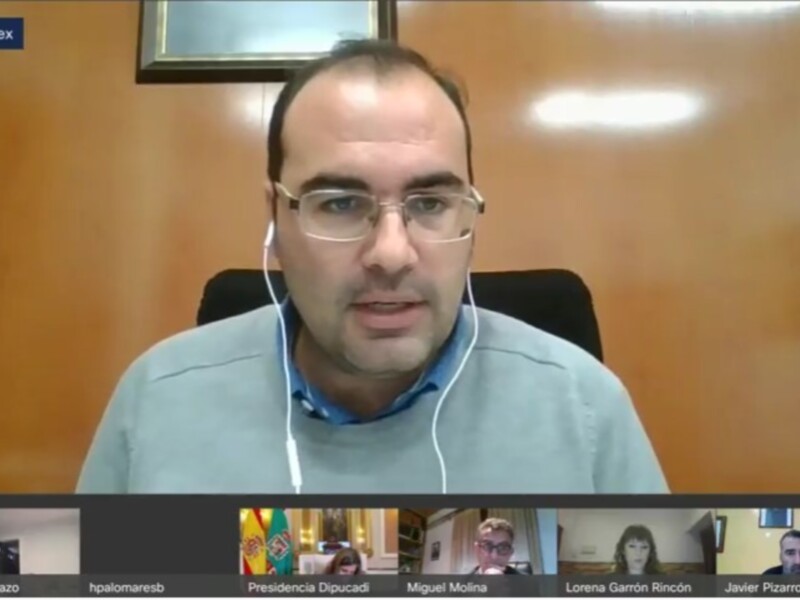 Un hombre con gafas y auriculares en una videoconferencia, con un fondo de pared naranja y una bandera española en la esquina inferior derecha.