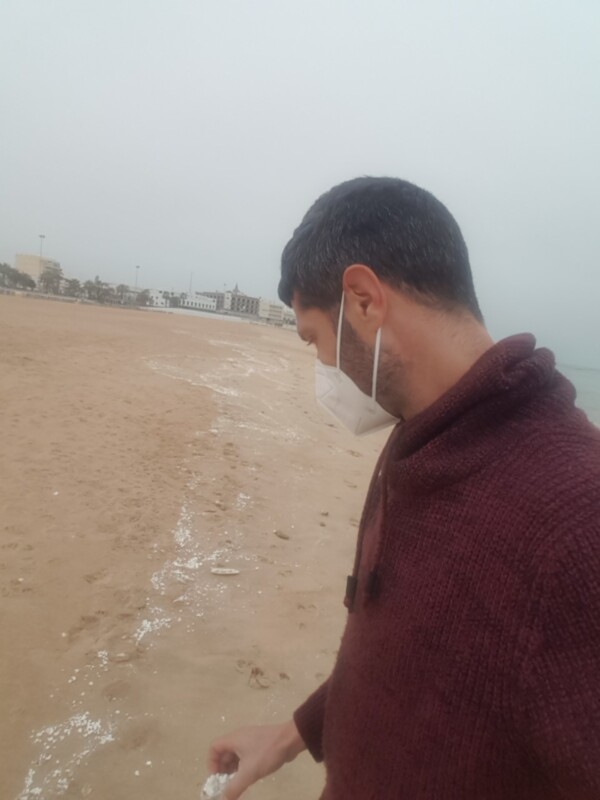 Un hombre con una máscara blanca y una gorra de lana se encuentra en una playa desierta, mirando hacia el mar en un día nublado.