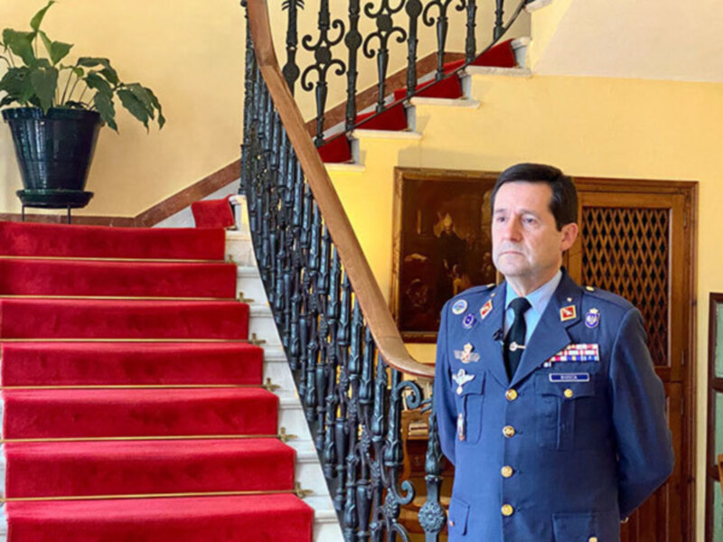 Un oficial militar en uniforme completo posa en una escalera con alfombra roja y barandilla de hierro forjado en un entorno formal.