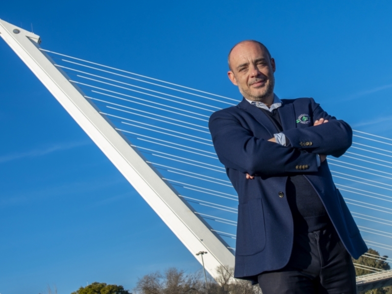 Un hombre en una chaqueta azul posa con las manos cruzadas frente a un puente moderno bajo un cielo azul claro.