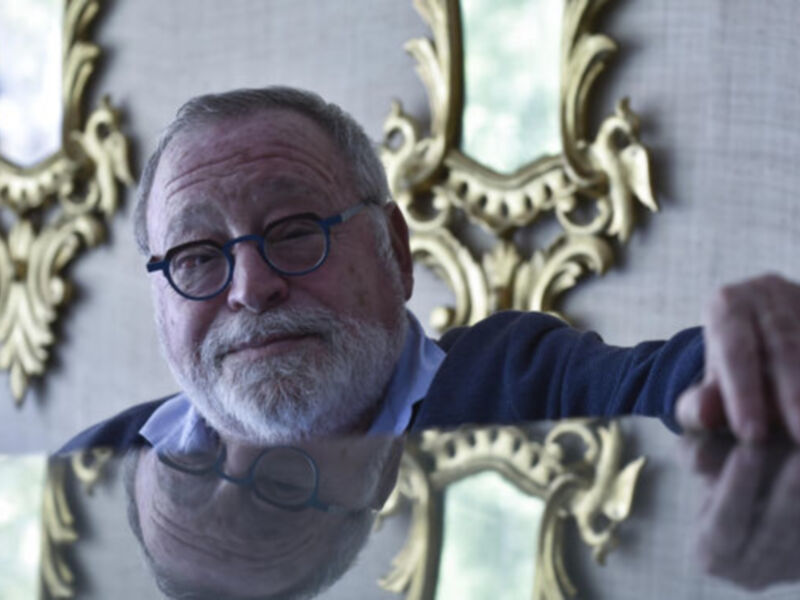 Retrato de un hombre con barba y gafas, apoyado en una mesa con espejos dorados en el fondo.