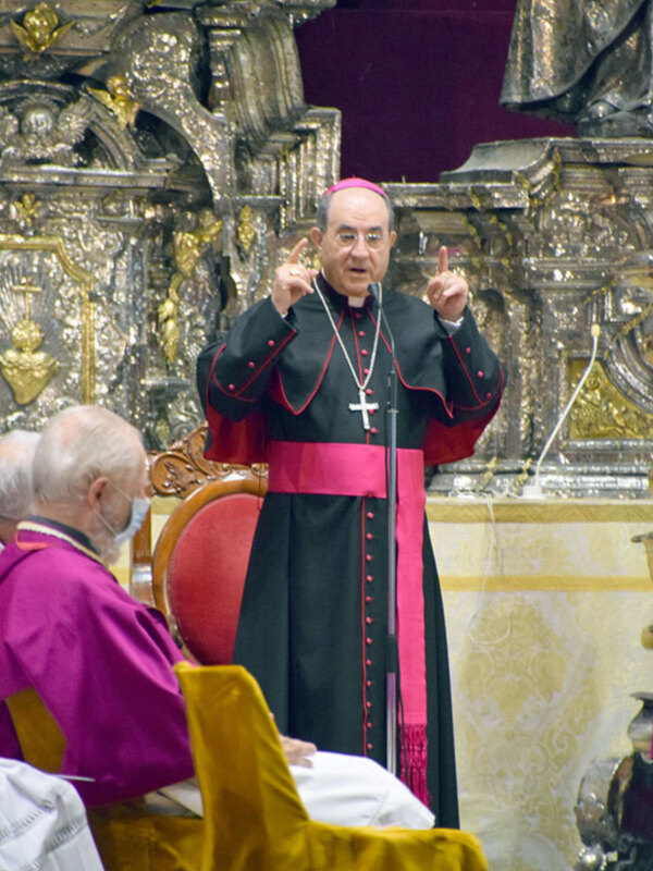 Cardenal con hábito de color negro y rojo, con una cruz en la peana, en un entorno de iglesia con detalles dorados y una estatua en el fondo.