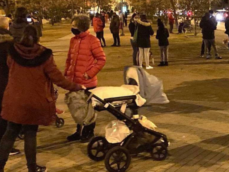 Gente caminando por la noche en una plaza, algunos con mascarillas y un cochecito de bebé.