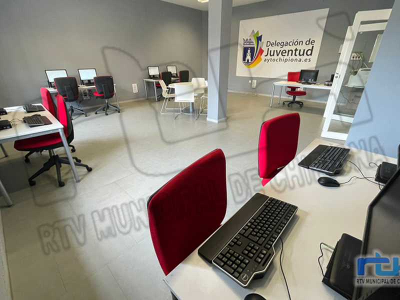 Oficina moderna con computadoras y sillas rojas, con un banner de la Delegación de Juventud en la pared.