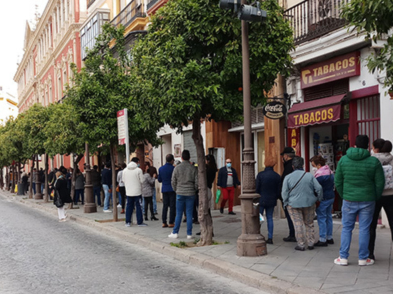 Línea de personas esperando en una calle con árboles y tiendas de tabaco en el fondo.