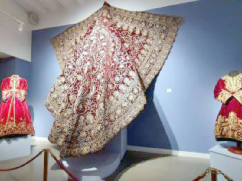 Exhibición de vestimentas tradicionales en un museo, destacando un manto ornado y dos trajes bordados en rojo y dorado.