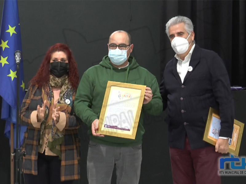 Un hombre sostiene un premio mientras una mujer y otro hombre lo aplauden en un evento formal con banderas de la UE y España en el fondo.