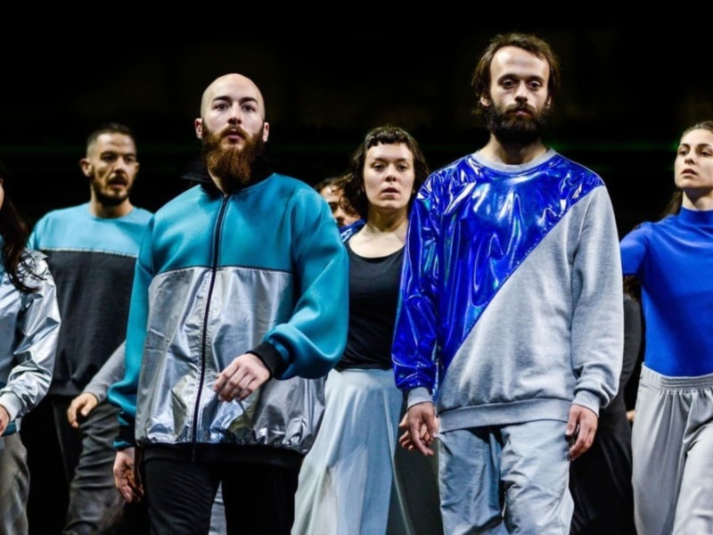 Un grupo de actores en trajes brillantes y uniformes caminan juntos en escena, creando una atmósfera teatral y colectiva.