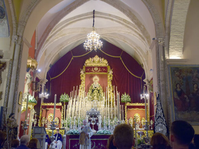 Interior de una iglesia con altares adornados con flores y velas, presidido por una imagen religiosa en un altar central, rodeado por fieles.