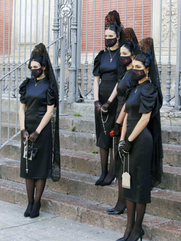 Damas vestidas con trajes tradicionales de negro y mantilla, posando en escaleras frente a una puerta de hierro forjado.