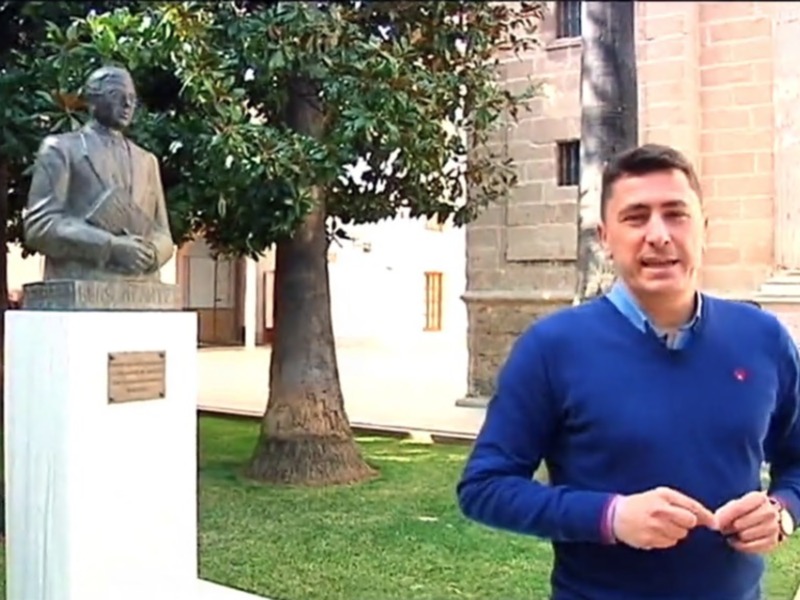 Un hombre habla frente a una estatua de un hombre en un jardín con edificios históricos al fondo.