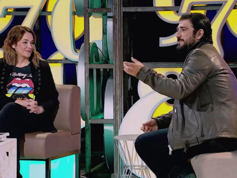 Una conversación en un programa de televisión, con un hombre y una mujer sentados en sillones frente a un fondo con letras grandes y colores brillantes.