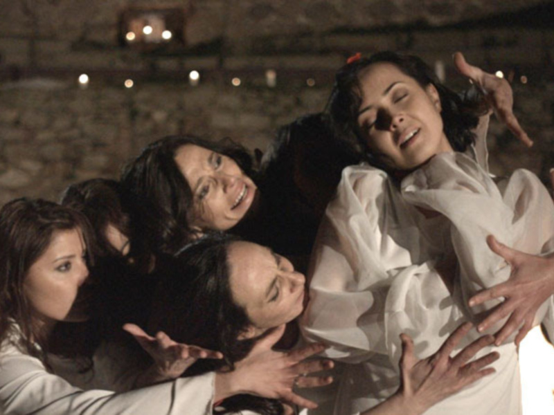 Un grupo de mujeres en blanco se abraza con ternura en un entorno sombrío, iluminado por velas y fuego, creando un ambiente emotivo y poderoso.