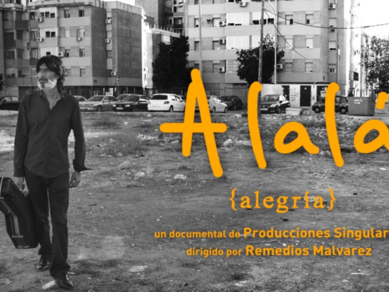 Un hombre con una guitarra camina por un área urbana desierta, con edificios de apartamentos en el fondo. Texto en el pie de foto: 'A la alegría, un documental de Producciones Singulares dirigido por Remedios Malvarez'.