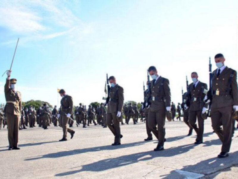 Grupo de soldados en uniforme militar, realizando una ceremonia o desfile, con máscaras faciales para prevenir el COVID-19. El cielo azul y la bandera nacional se destacan en el fondo.