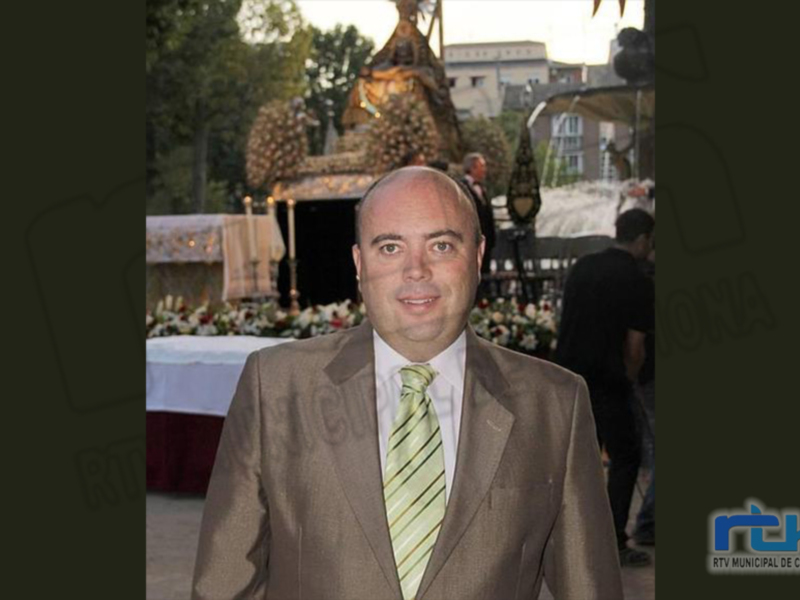 Hombre en traje y corbata en un evento público con decoración floral y estructuras ornamentales al fondo.