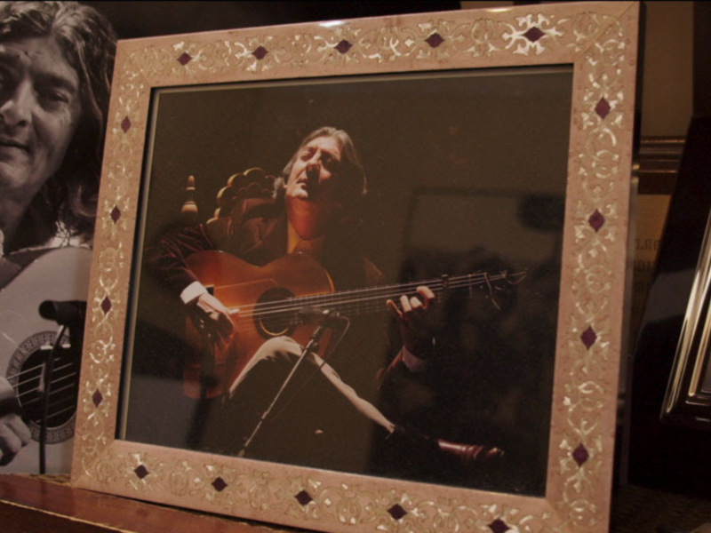 Fotografía de un músico tocando la guitarra, rodeado por objetos relacionados con su música, como una guitarra y un marco con una imagen de él mismo.