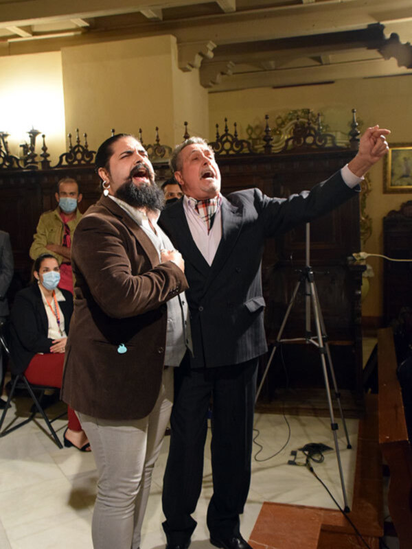 Dos hombres en trajes cantando en un entorno formal con una audiencia en el fondo.