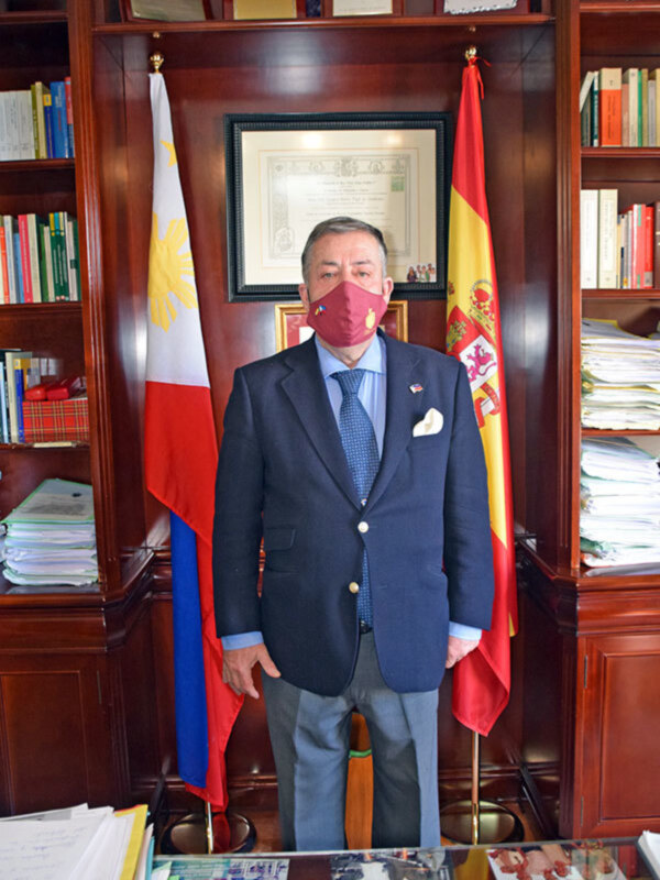 Un hombre en traje con máscara, detrás de banderas filipinas y españolas, en un entorno formal con estanterías de libros y documentos.