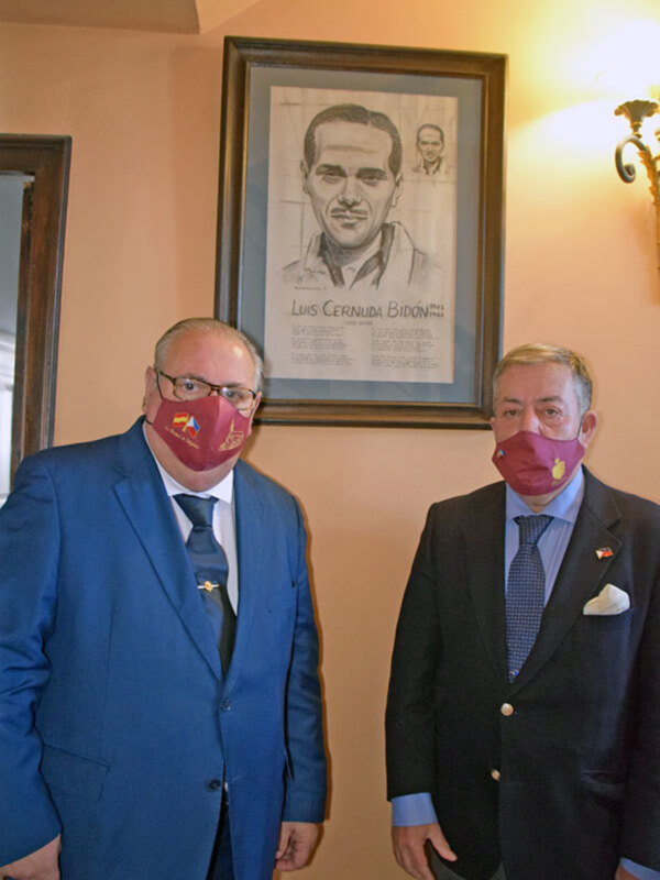 Dos hombres en trajes y mascarillas rojas posan frente a un retrato de Luis Cernuda Bidón en un ambiente formal.