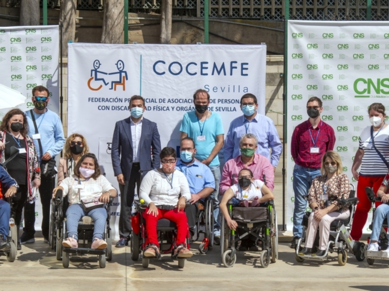 Fotografía de una reunión de personas con discapacidad física y organizaciones de Sevilla, con un grupo de personas en sillas de ruedas y otros miembros de la comunidad, frente a una pancarta de COCEMFE y CNS.