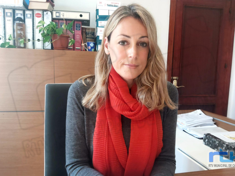 Una mujer con cabello largo y rubio, vistiendo una camiseta gris y un pañuelo rojo, sentada en una oficina con una puerta de madera y un escritorio con papeles. El entorno muestra un ambiente laboral formal.