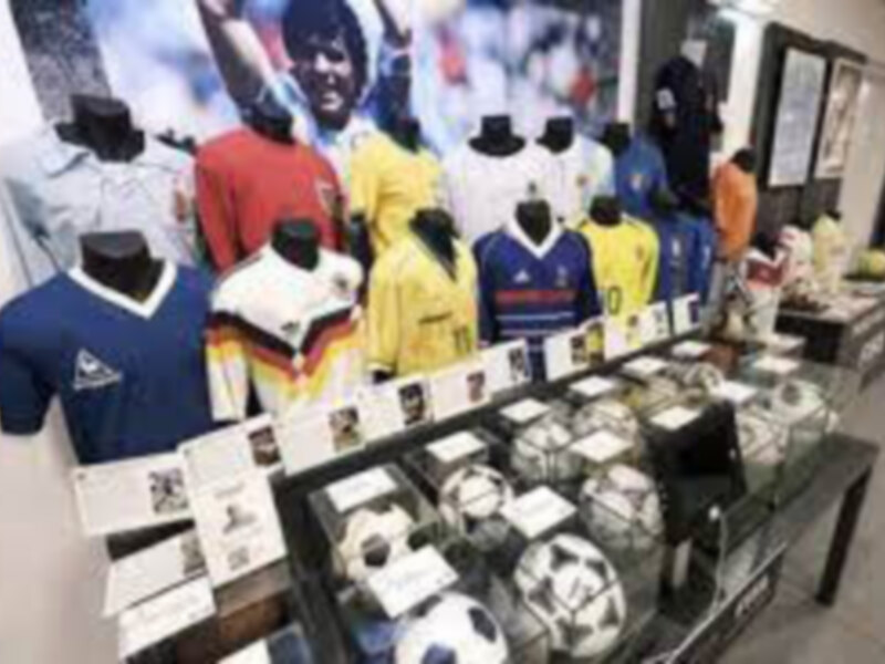 Se muestra una colección de camisetas deportivas en diferentes colores y estilos, junto con varios accesorios como medallas y etiquetas. El fondo es un espacio interior bien iluminado, probablemente una tienda o exposición de ropa deportiva.
