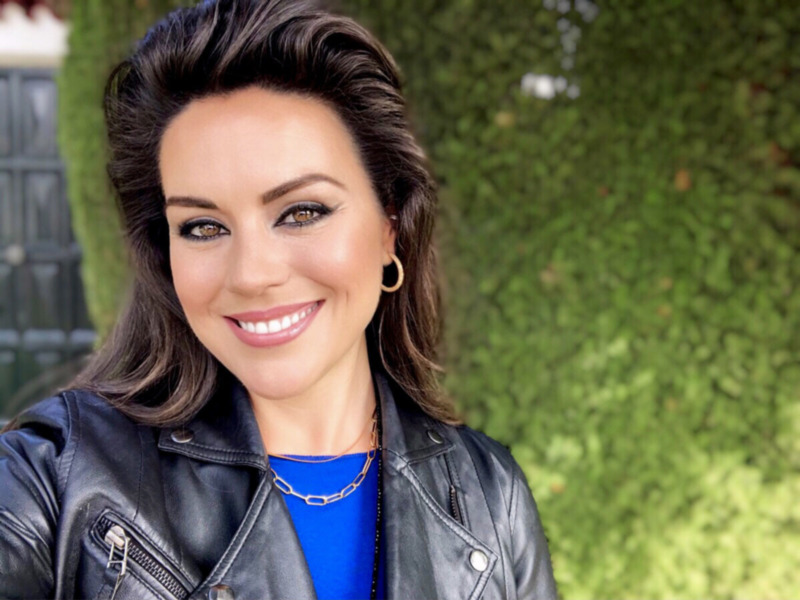 Elegante mujer con cabello oscuro y maquillaje suave, vestida con una chaqueta de cuero y una camisa azul. Fondo verde con detalles de luz.
