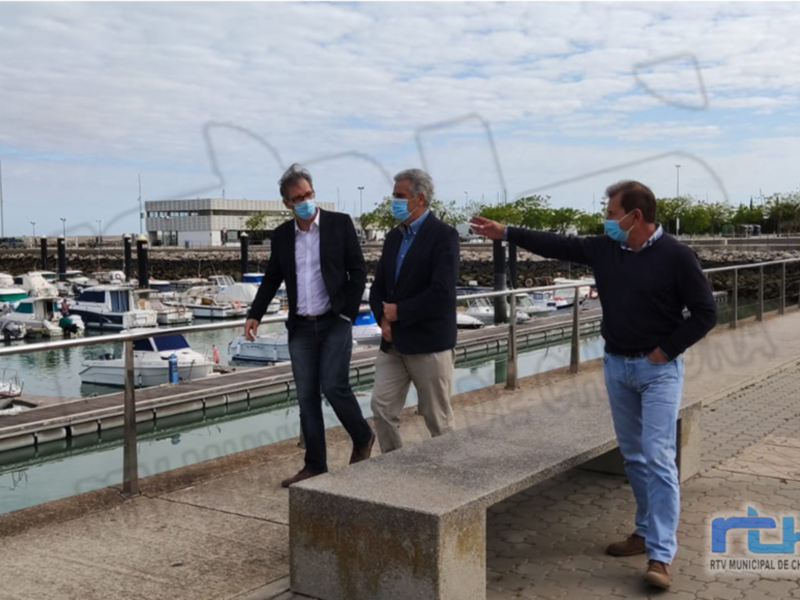 Tres hombres discuten en un muelle junto al puerto, con barcos y un edificio al fondo.