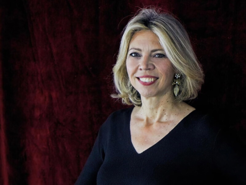 Elegante retrato de una mujer con cabello largo y rubio, vestida con un suéter negro y joyas en el cuello, frente a un fondo rojo.