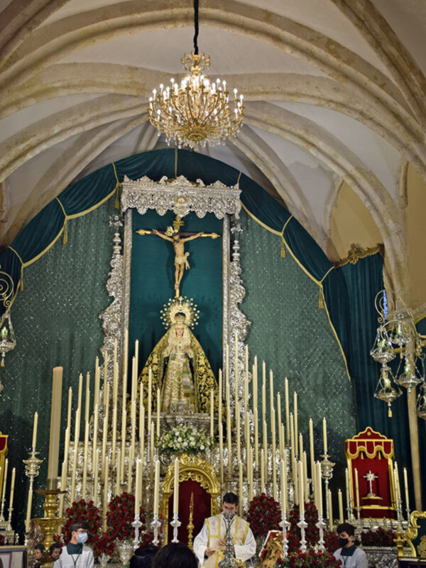 Interior de una iglesia con altar ornado, imagen de la Virgen y un sacerdote oficiando una misa.