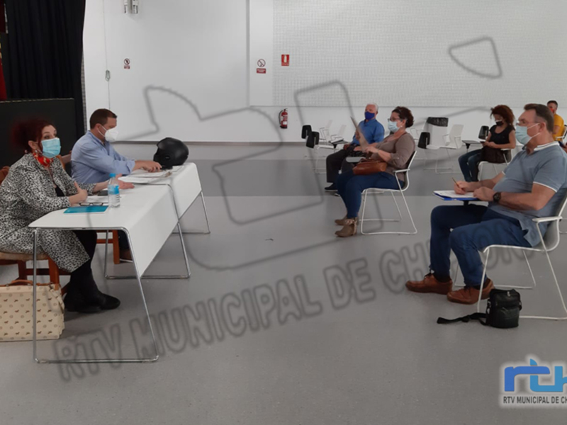 Reunión de trabajo en una sala de conferencias con personas sentadas en mesas, todos con mascarillas y documentos en sus manos.