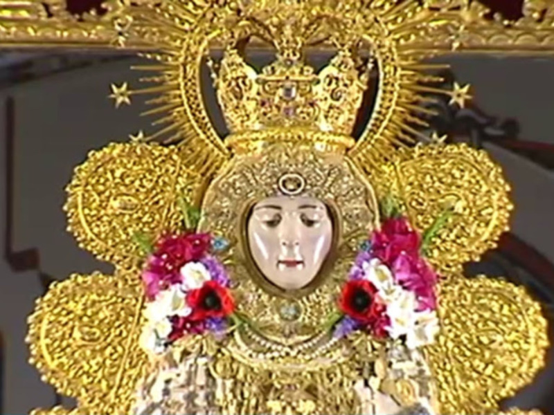 Una imagen de la Virgen María vestida con una corona dorada y flores en su frente, en un contexto religioso o cultural.