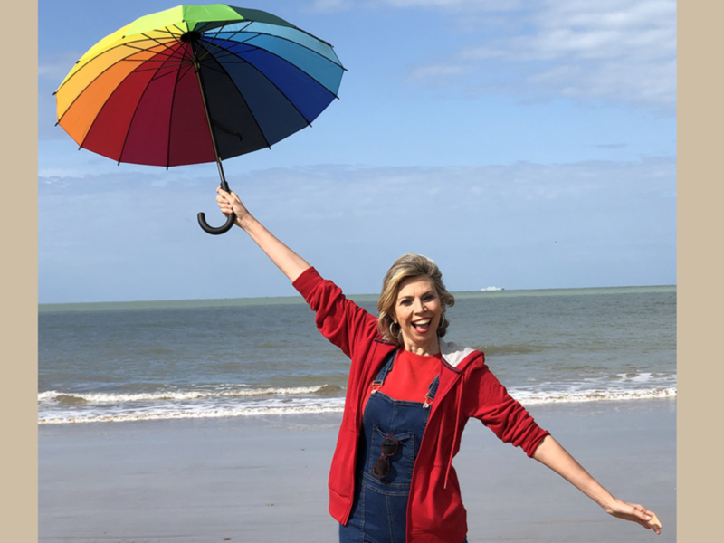 Una mujer sonríe en la playa sosteniendo un paraguas de colores arcoíris, con el mar y el cielo de fondo.