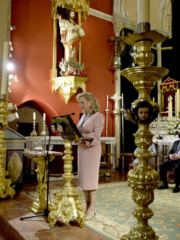 Se observa a una mujer vestida con un elegante abrigo rosa, posiblemente en una ceremonia religiosa o de honor. La escena se desarrolla en un entorno decorado con elementos dorados y una estatua de Cristo en el fondo. La mujer está sosteniendo un libro o un objeto similar, lo que sugiere una actitud de respeto o reverencia. La atmósfera es formal y solemnemente decorada, con detalles que indican un ambiente de alto estatus o celebración importante.
