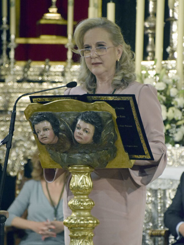 Una mujer con cabello rubio y vestida de rosa sostiene una imagen dorada que muestra a un niño y a una mujer. La escena parece ser en un evento formal, con luces doradas y un fondo de velas.