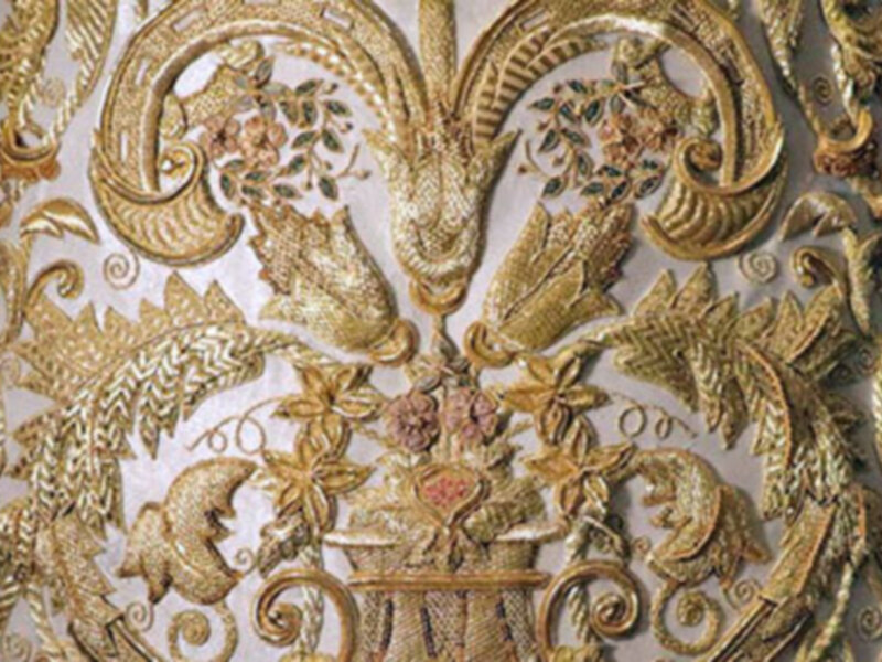 Detalle de un bordado dorado con motivos florales y serpientes, con un estilo ornamental y sofisticado.