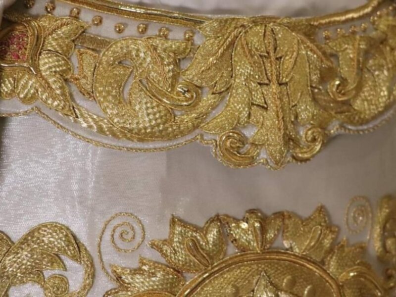 Detalle de bordado dorado en un tejido blanco, con patrones florales y volutas.