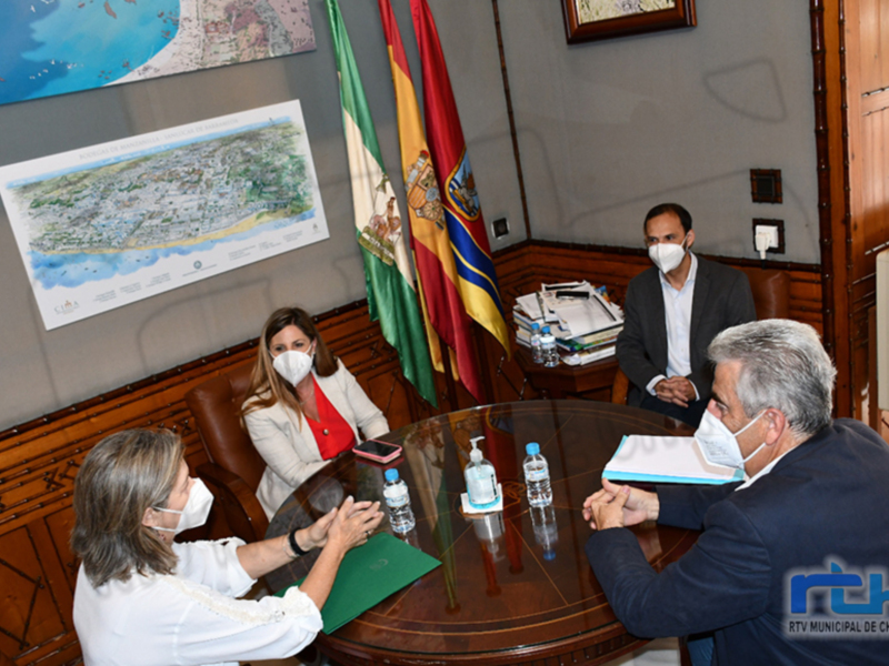 Tres personas sentadas en una sala de reuniones, con una bandera española y un mapa en el fondo. Las personas están usando máscaras faciales y hay documentos y botellas de agua en la mesa. La imagen parece ser de una reunión oficial o de trabajo.