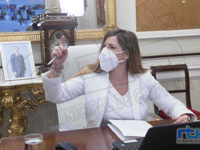 Mujer en traje blanco y mascarilla, sentada en un escritorio con un ordenador, apuntando con un bolígrafo.