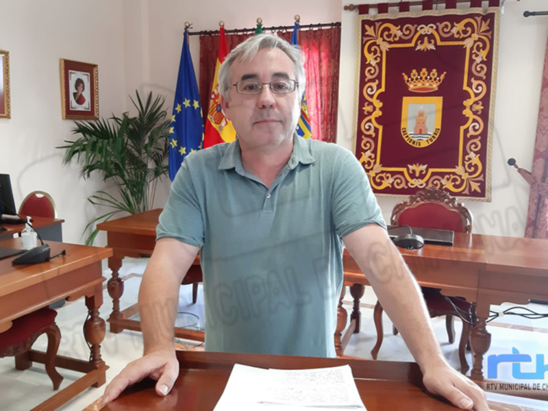 Un hombre en una oficina municipal, con una bandera española y una bandera europea en el fondo, posando para una foto.